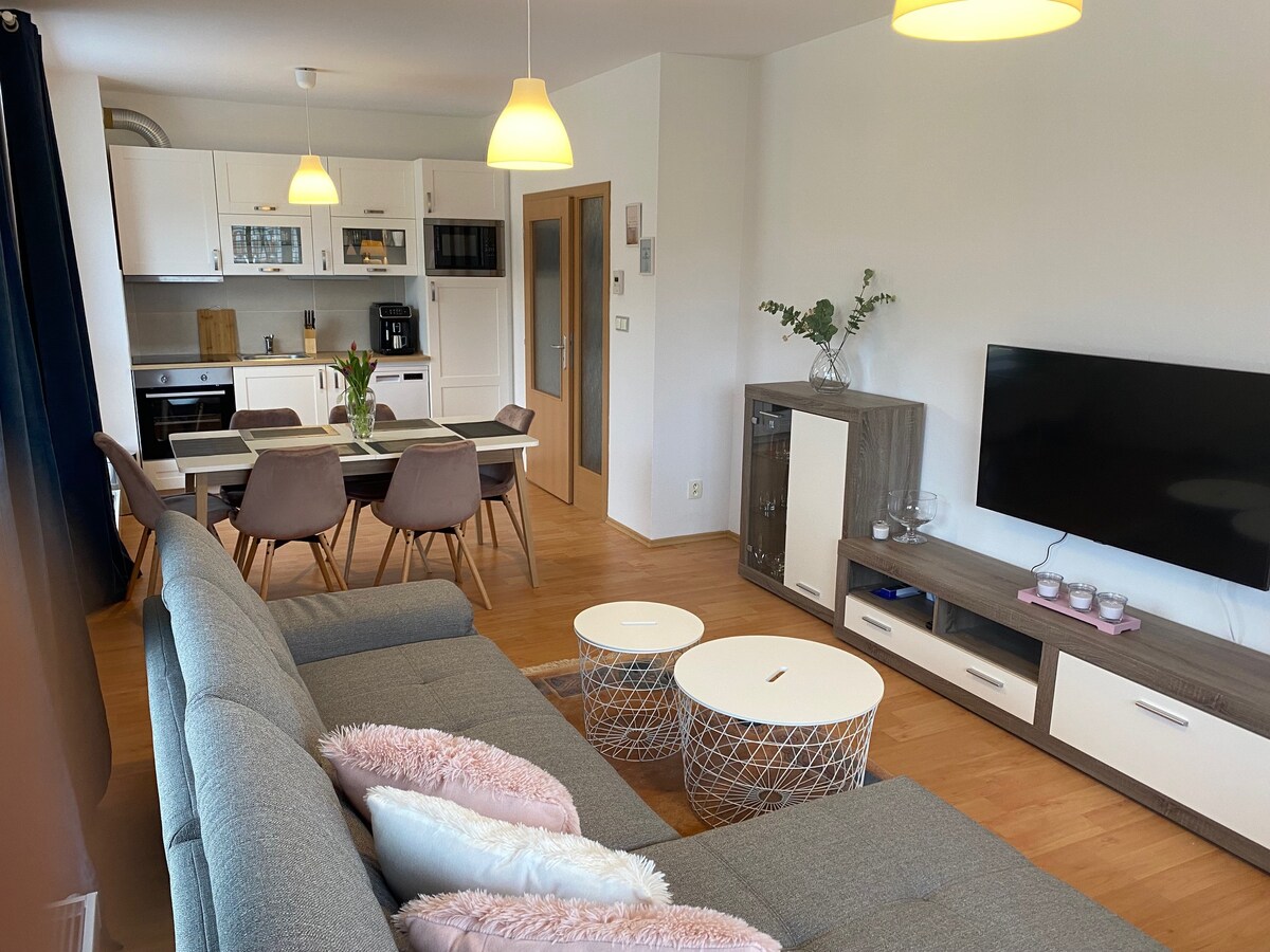 The open-plan living space features a modern kitchen and dining area with a large table surrounded by stylish chairs. A comfortable gray sofa is positioned in front of a wall-mounted TV, with two circular coffee tables placed nearby. Soft lighting is provided by hanging fixtures.