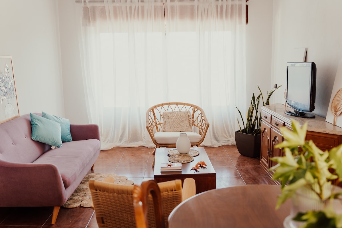 A cozy living area is furnished with a lavender sofa and a rattan chair, providing comfortable seating. A wooden coffee table rests in the center, decorated with a vase. Natural light filters through sheer curtains, illuminating the space.