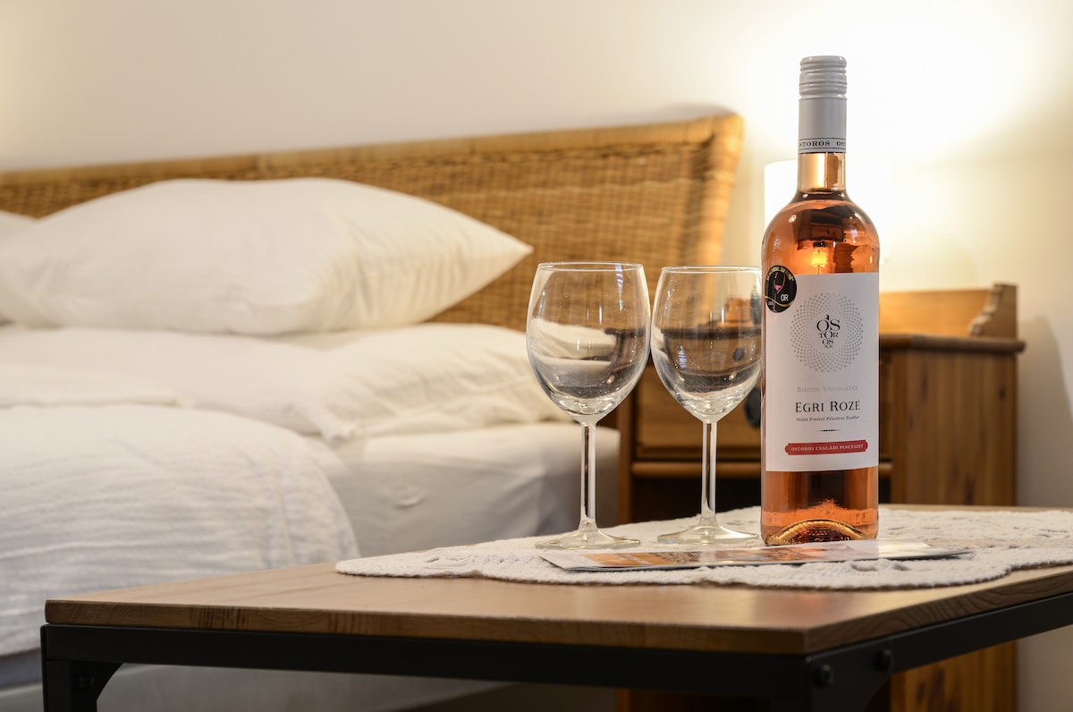 A wooden table features a bottle of Egri Rosé wine alongside two empty wine glasses. In the background, a spacious bed awaits with crisp white linens, blending a sense of relaxation and comfort in a serene setting.