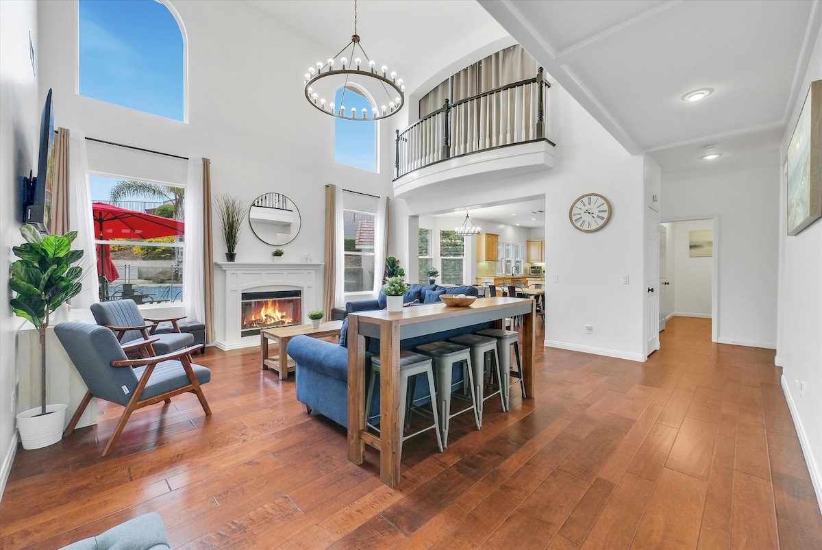 A bright, open living space features high ceilings and a large chandelier above a wooden dining table. A cozy fireplace is visible, complemented by large windows that allow natural light to brighten the room. Comfortable seating is arranged, creating an inviting atmosphere.