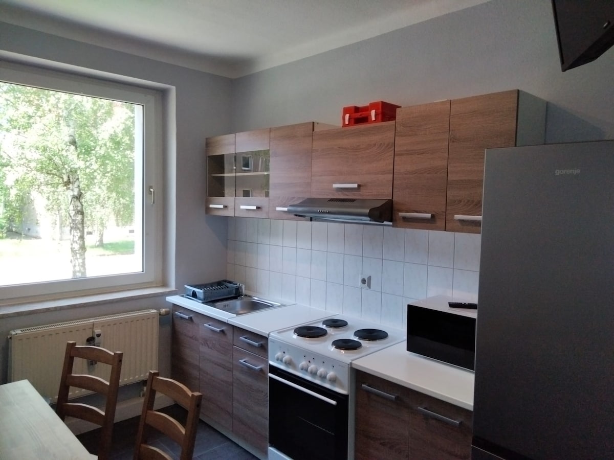 A kitchen area is presented, featuring wooden cabinetry and modern appliances. The space includes a stove, oven, and microwave, with a window providing natural light and a view of greenery outside. A dining table with several chairs is visible.