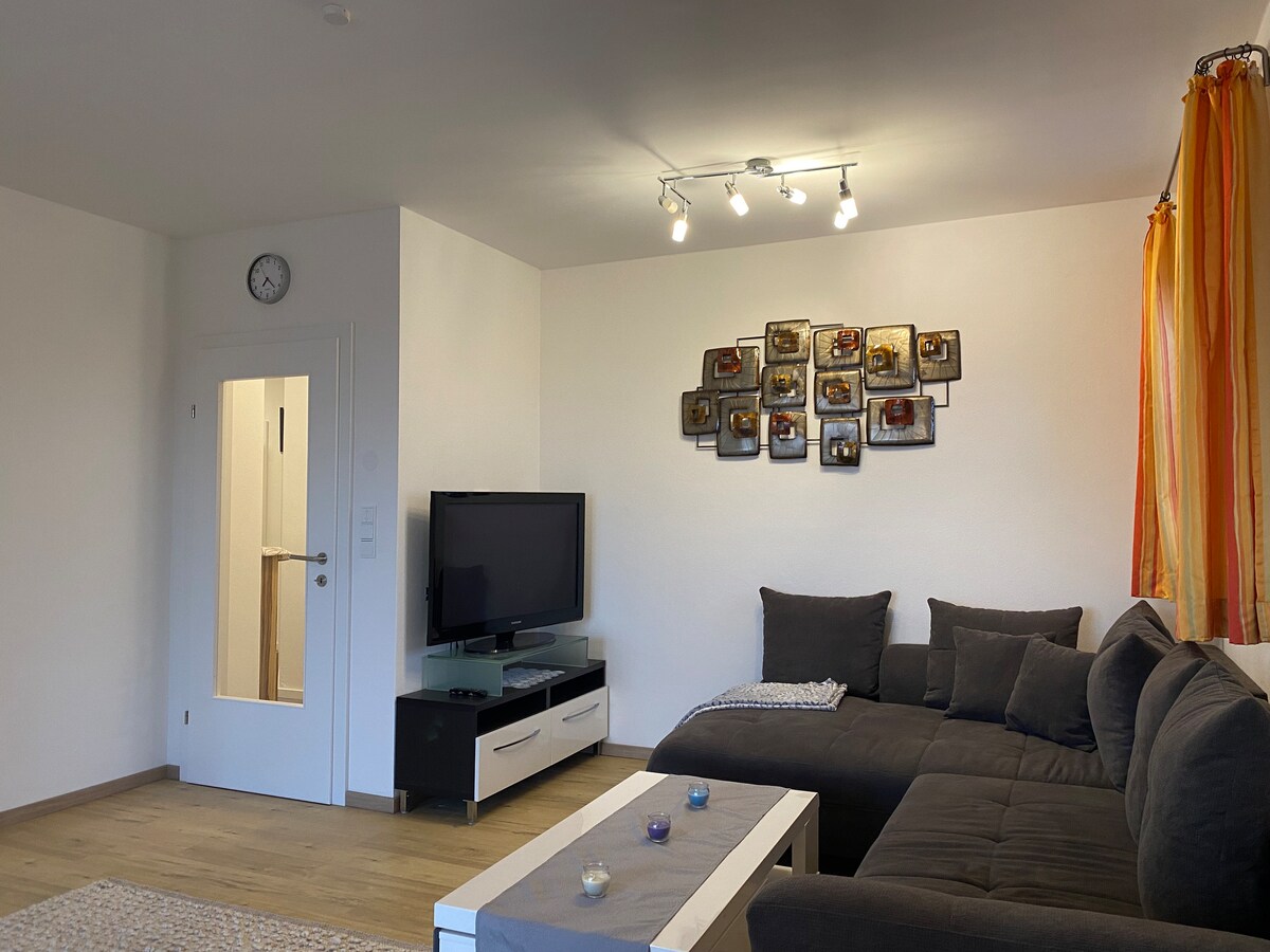 A spacious living area features a dark sectional sofa arranged around a coffee table. A flat-screen television is positioned on a modern stand. A collection of framed photographs decorates the wall, and natural light is invited through curtains on one side. A wall clock is also visible.