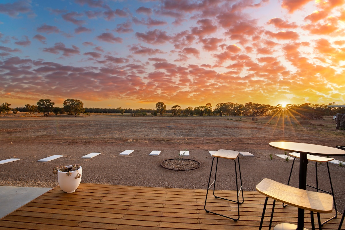 A tranquil outdoor deck area is presented, featuring modern furniture that includes two wooden tables and a planter. The horizon showcases a colorful sunset over the distant fields, with clouds illuminated in shades of pink and orange, and soft rays of sunlight breaking through.