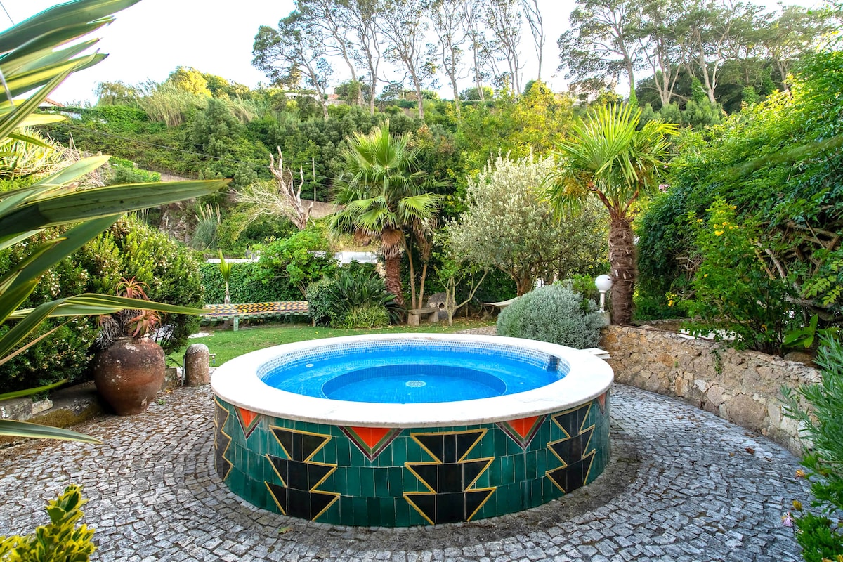 A round hot tub is surrounded by a lush garden with various plants and trees. The tub features colorful tile work, while the natural stone path leads to a green space with lounge chairs, creating a peaceful outdoor retreat.