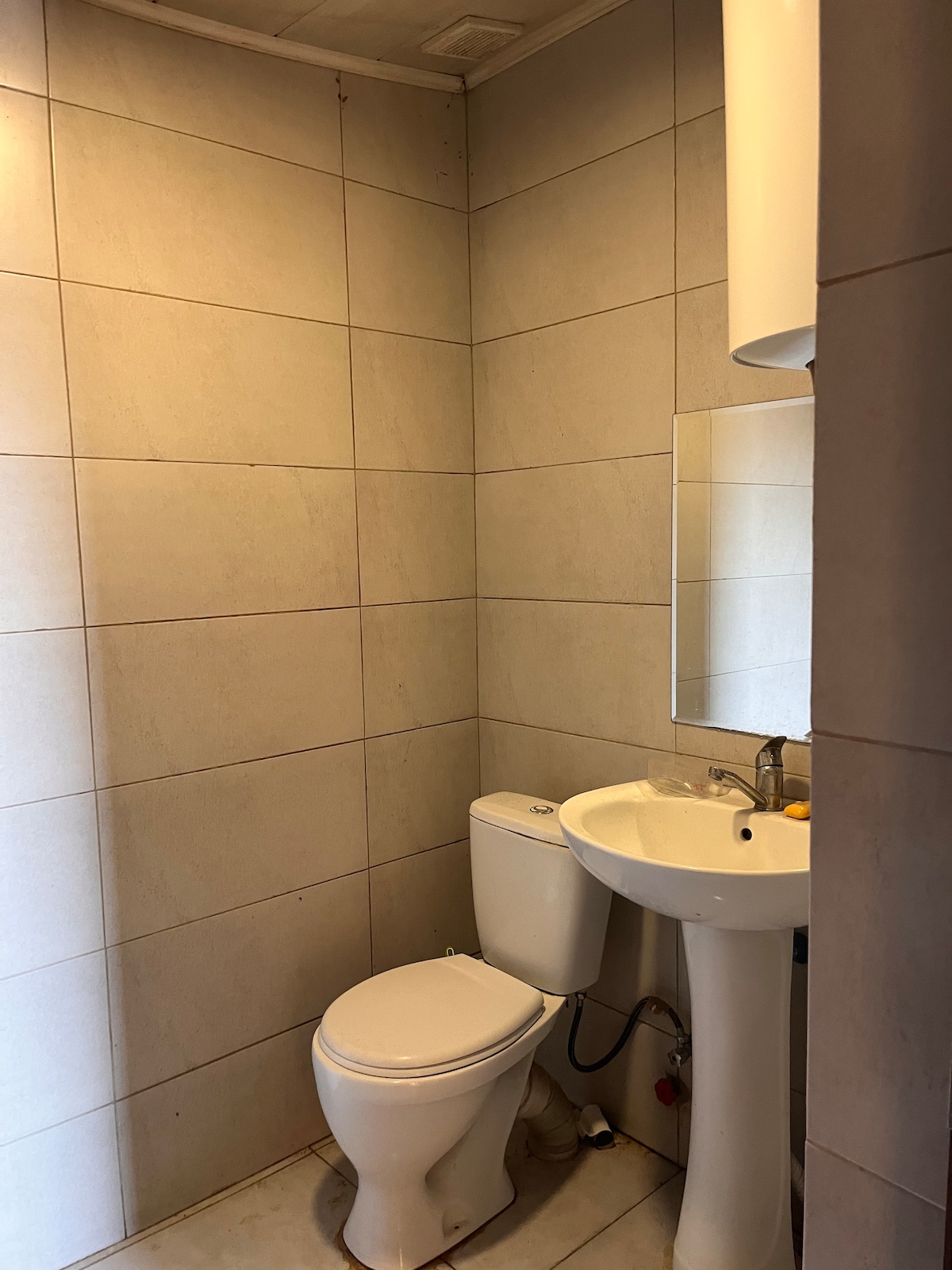 A compact bathroom is presented, featuring light-colored tiles that cover the walls. A modern toilet is positioned alongside a pedestal sink with a mirror above. A small shelf holds toiletries, contributing to the functional layout of the space.