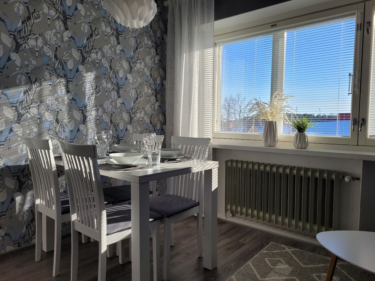 A bright dining area features a white table set for four, with glassware and dishes neatly arranged. Light streams through a large window adorned with sheer curtains, revealing a view of the skyline. Floral wallpaper adds a touch of elegance to the space.