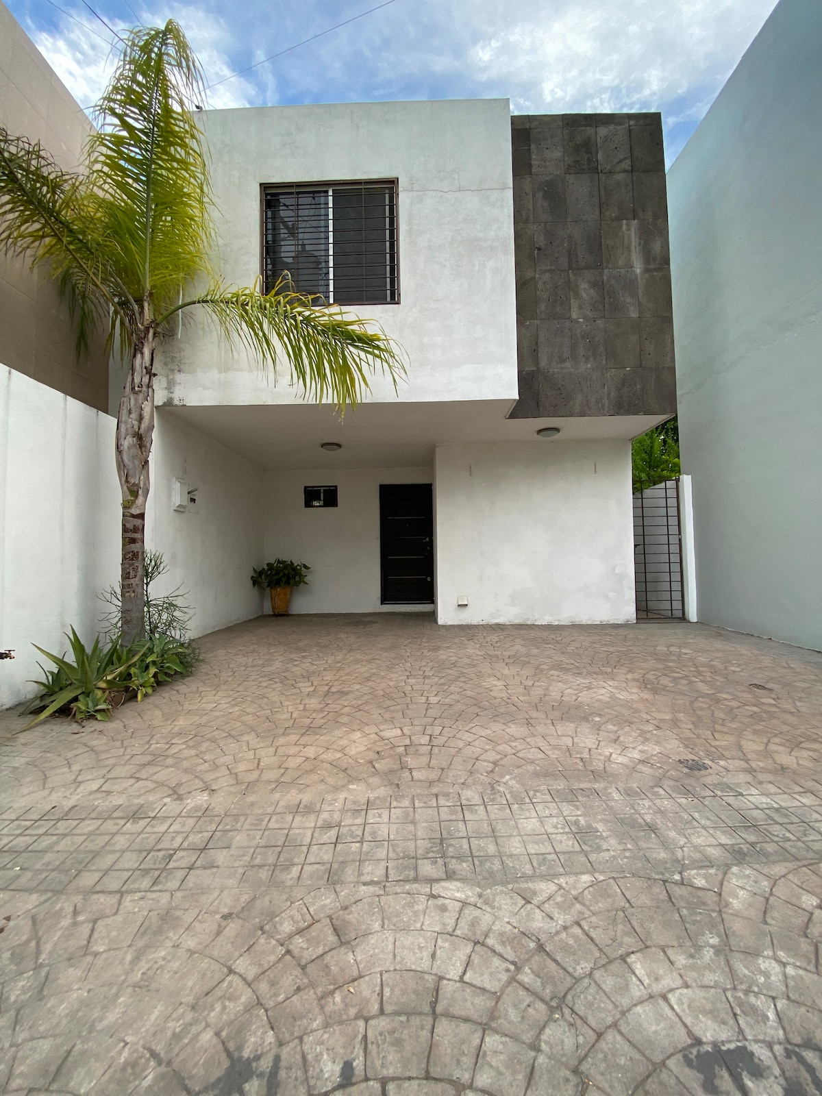 The exterior of a modern two-story house is shown, featuring a textured concrete facade and a well-maintained driveway. A palm tree and potted plant add greenery to the scene. Large windows provide natural light to the upper level, contributing to the home's inviting appearance.