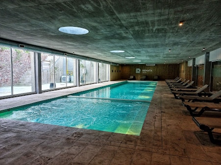 An indoor swimming pool is highlighted with large glass windows allowing natural light to fill the space. Comfortable lounge chairs are arranged alongside the pool. Textured walls and a smooth ceiling provide a modern aesthetic, creating a relaxing environment.
