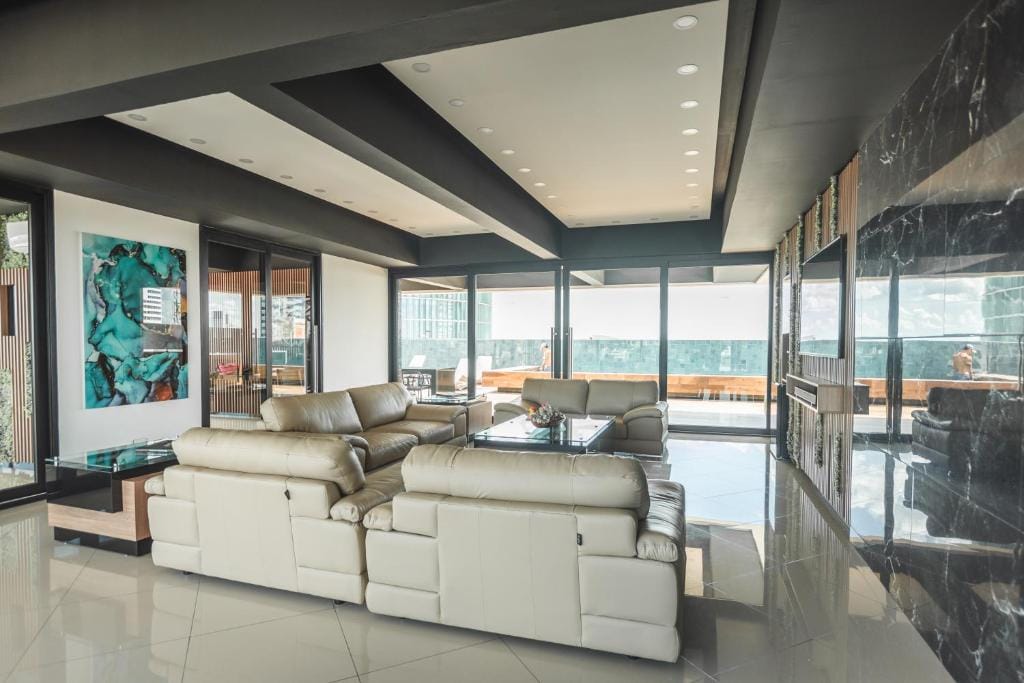 An open living area is showcased, featuring a large L-shaped cream sectional sofa and glass coffee table. Floor-to-ceiling windows provide a view of the surrounding cityscape, while the ceiling design offers a modern touch. Natural light fills the space, enhancing its spacious layout.