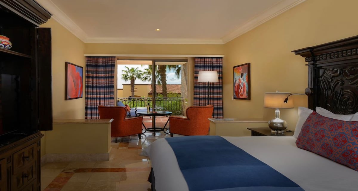 A Junior Suite is visible, featuring a spacious bed with a blue throw and decorative pillows. A seating area is arranged with two orange chairs and a small table, while a balcony offers views of palm trees. Artwork adorns the walls, and large windows provide natural light.