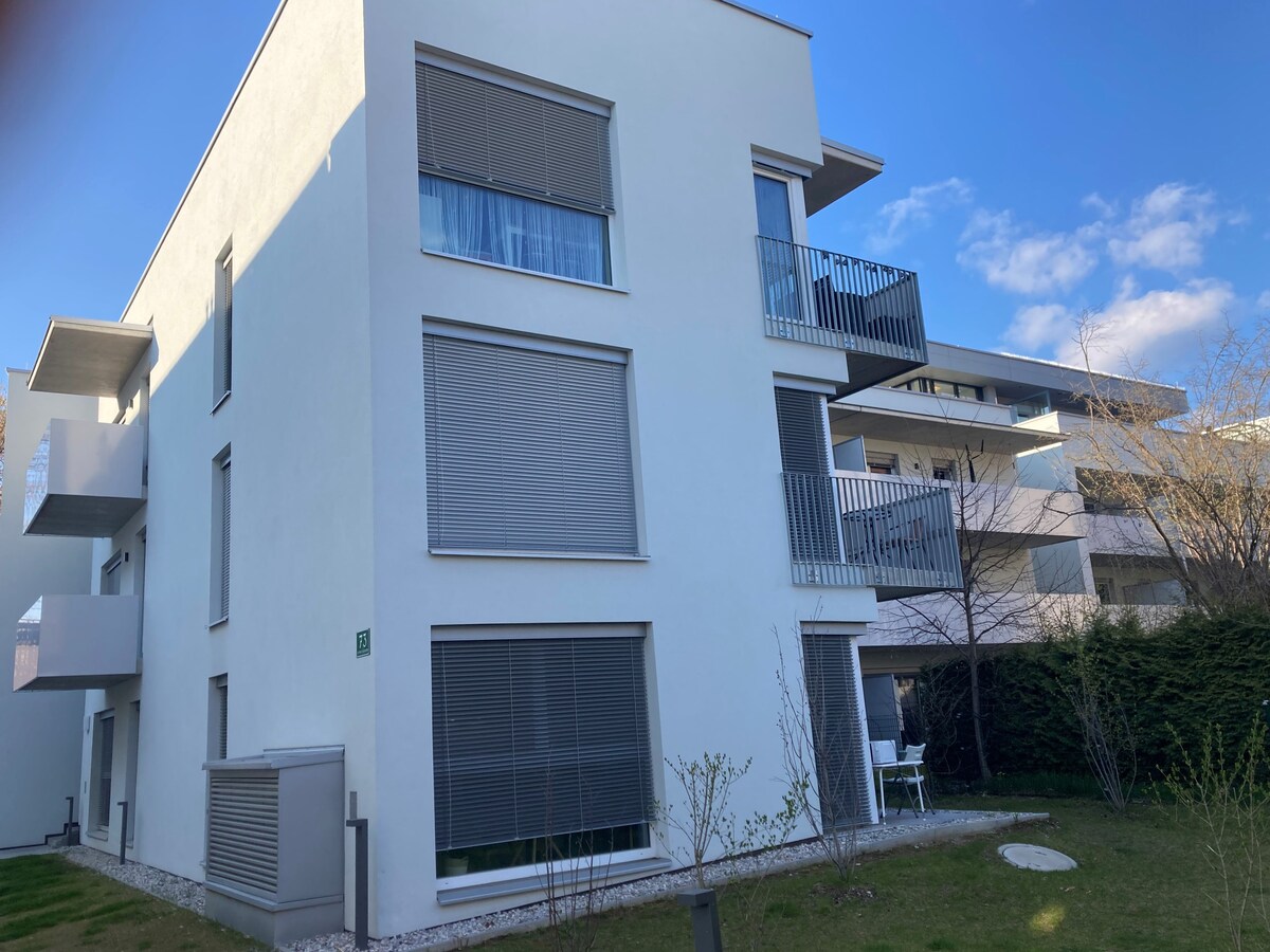 A modern building is shown from the exterior, featuring multiple windows with sleek horizontal blinds. Balconies are visible on the upper floors, enhancing the structure's contemporary design. A manicured garden with small shrubs and a lawn surrounds the building, contributing to a serene atmosphere.