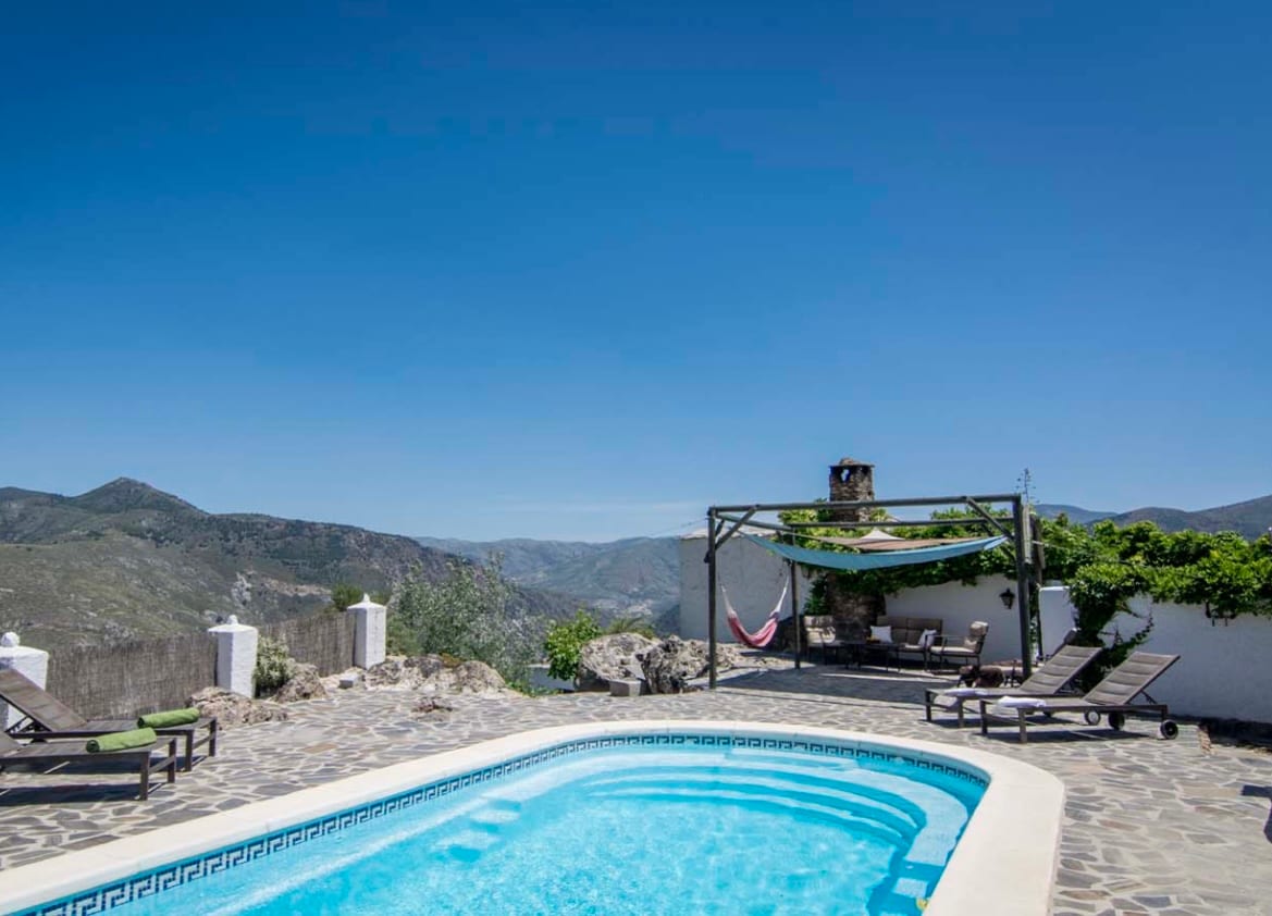A private swimming pool is surrounded by stone decking and lounge chairs. An outdoor seating area with a hammock is situated under a pergola, offering views of the mountainous landscape in the background. Clear blue skies enhance the serene atmosphere.