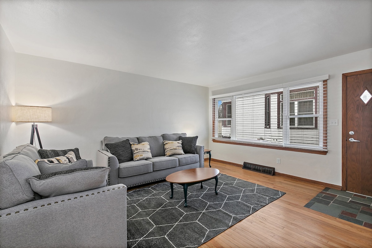 A cozy living area is furnished with a gray sofa and armchair, complemented by accent pillows. A round coffee table rests on a geometric rug, while natural light pours in through a large window with blinds. A soft glow is provided by a lamp in the corner.