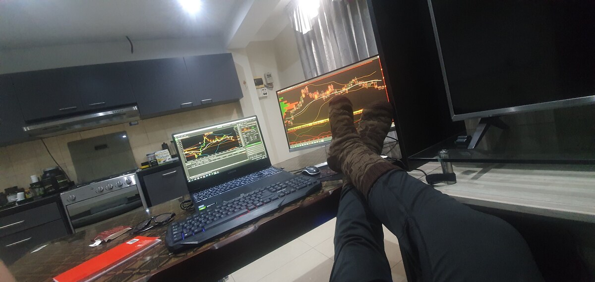 A modern workspace is depicted, featuring a sleek kitchen area with dark cabinetry and stainless steel appliances. Dual monitors display financial graphs. A desk with a keyboard and a comfortable chair are positioned nearby. A person's feet, adorned in cozy socks, rest on the desk.