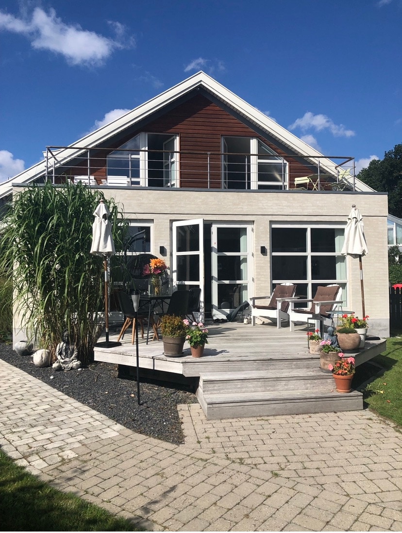 The two-story house features a combination of white and wooden siding, surrounded by green foliage. A welcoming wooden deck leads to doors framed by large windows. Potted plants and chairs create a cozy outdoor space, while a balcony extends from the upper level.