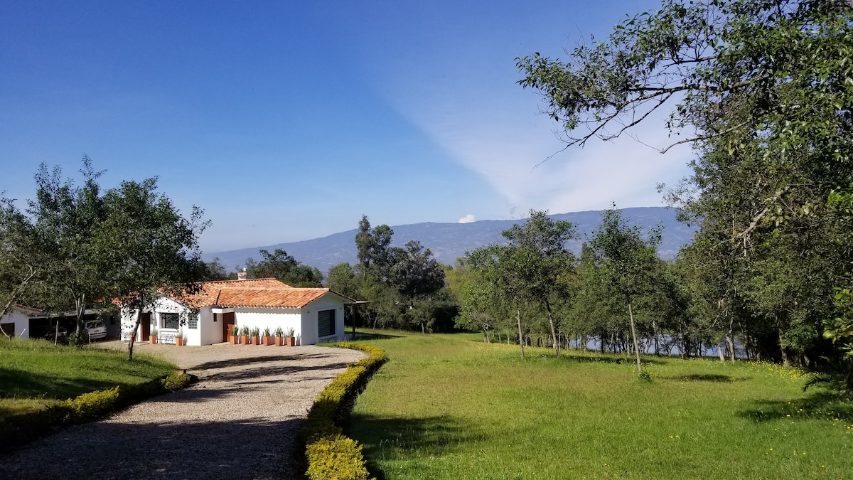 A gravel pathway leads to a white building with a terra-cotta roof, surrounded by lush green grass and trees. In the background, distant mountains are visible beneath a clear blue sky.