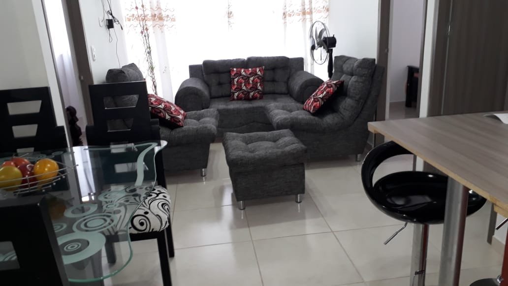 A modern living area features a large gray sectional sofa adorned with patterned throw pillows. A small gray ottoman is positioned in front of the sofa. A glass dining table with black and white patterned chairs is visible, along with a bowl of colorful fruits on the table.