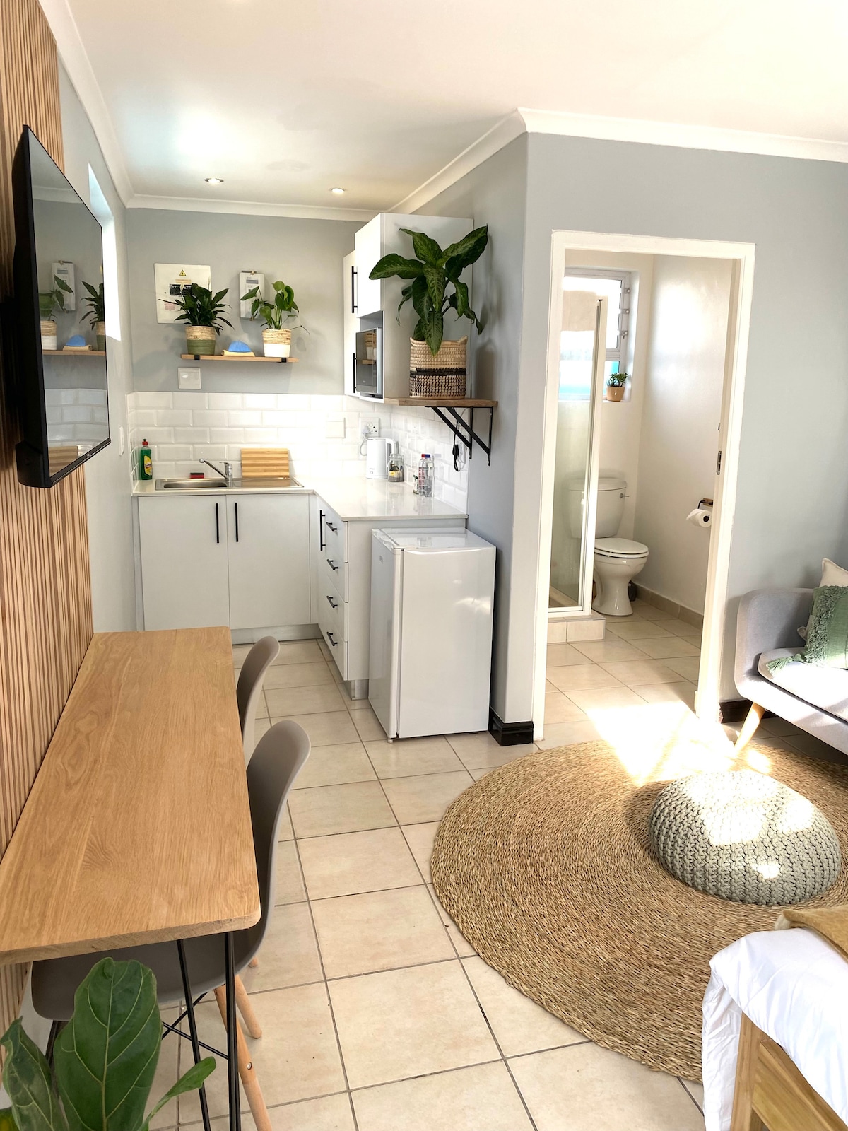 A bright and modern studio apartment is showcased with a compact kitchen featuring a refrigerator and countertop. A dining area with two chairs is positioned alongside the kitchen. A comfortable seating area is visible, with a decorative rug enhancing the ambiance.