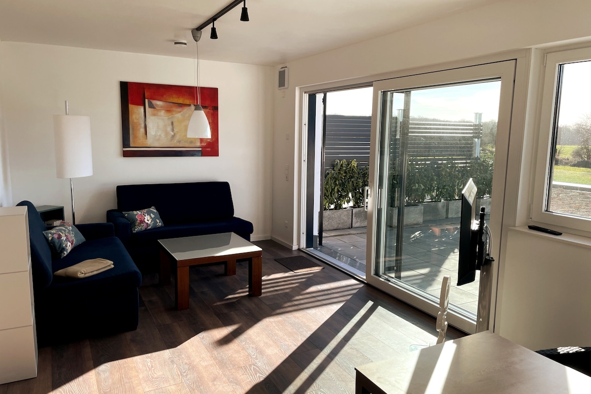 A modern living space features a blue sofa with decorative pillows, a wooden coffee table, and an adjacent dining area. Large glass doors allow natural light to fill the room and provide access to the outdoor terrace, surrounded by greenery.