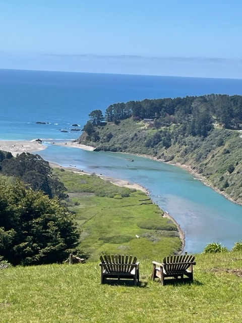 Two rustic wooden chairs are positioned on a grassy hill, overlooking a winding river that flows into the ocean. Lush greenery surrounds the area, and the clear blue waters of the ocean meet the sky in the distance, creating a serene coastal view.