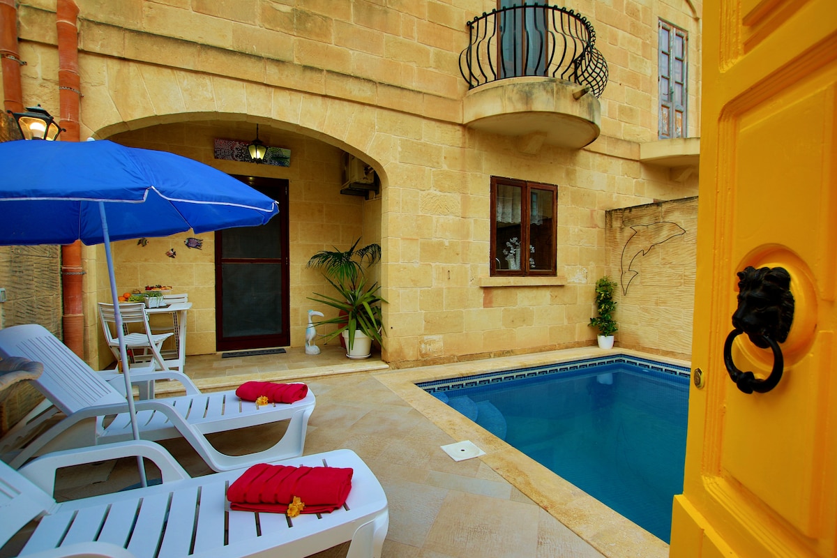 A private outdoor space is visible, featuring a clear swimming pool surrounded by sun loungers. A vibrant yellow door frames the entrance, while an umbrella provides shade. Tropical plants enhance the setting against stone walls, creating a calm retreat atmosphere.