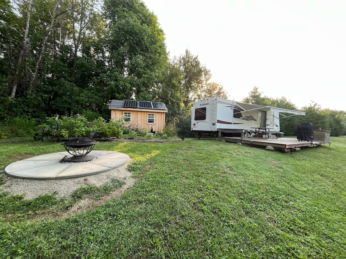 A rustic wooden cabin, equipped with solar panels, is positioned beside a spacious RV. A circular fire pit surrounded by gravel is situated in the grassy area, complemented by lush greenery and trees in the background.