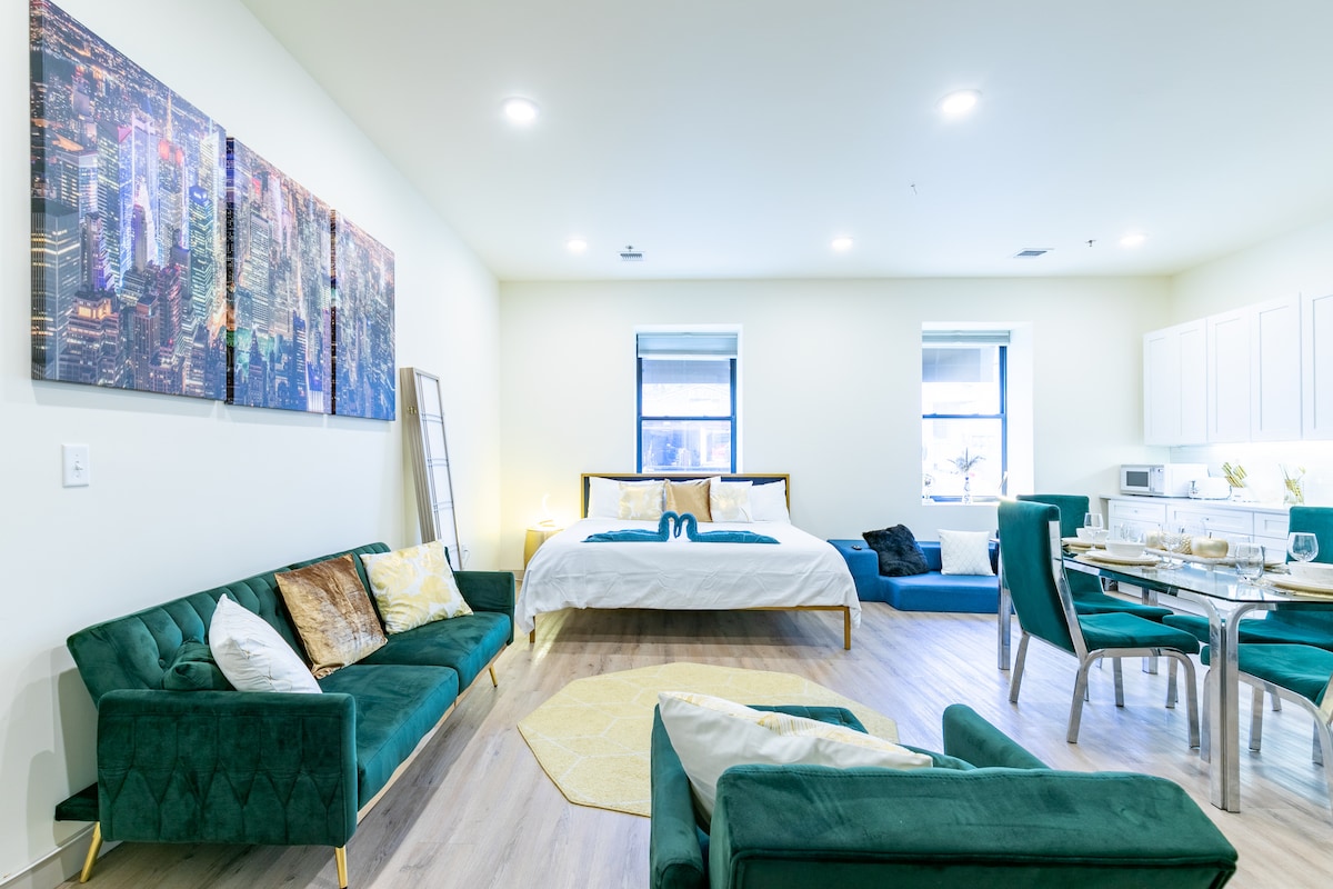 A spacious studio apartment features a large king bed with a white duvet, flanked by two windows. A green sofa set is arranged to create a cozy seating area, and a dining table for four is visible, complementing the open layout. Modern kitchen appliances are located in the background.