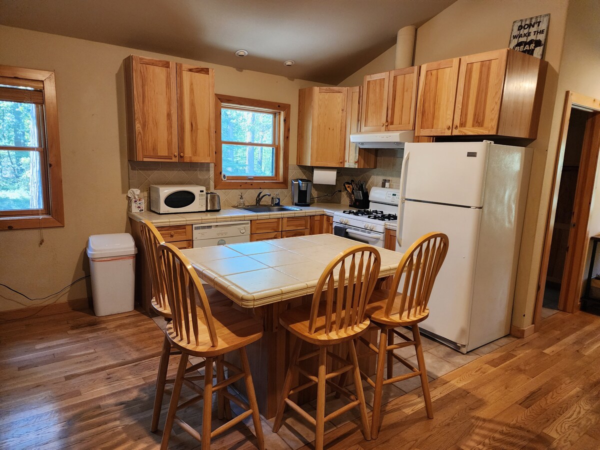 A functional kitchen features wooden cabinetry, light-toned countertops, and essential appliances. A table with four chairs occupies the center, while natural light streams through two windows. Additional items visible include a microwave, refrigerator, and stove, contributing to a practical cooking space.