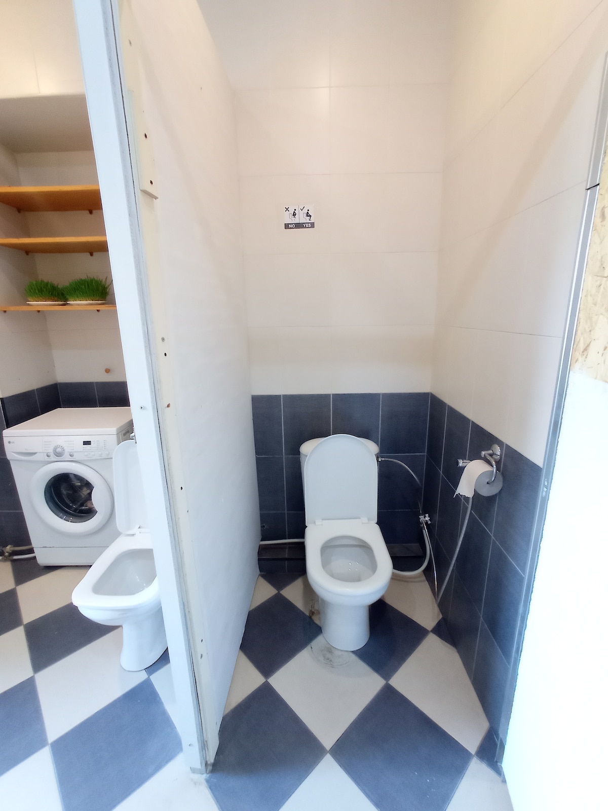 A functional bathroom space is presented with two toilets and a washing machine. Light-colored walls contrast with dark tiled flooring, enhancing the sense of cleanliness. Shelving is visible above the machine, offering practical storage for toiletries or other essentials.