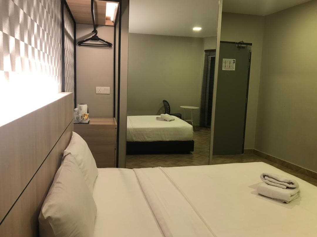 A warm room is seen featuring a spacious bed neatly dressed with white linens. A mirror reflects a second bed in the background. A wardrobe with hangers and a small desk fan are visible, while soft lighting enhances the overall ambiance.