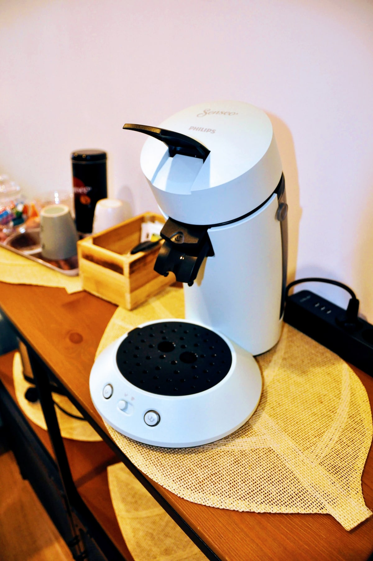 A coffee machine is positioned on a wooden surface, accompanied by a small wooden tray containing coffee pods. The machine features a sleek white design, with a black handle and a circular base that includes a button panel.