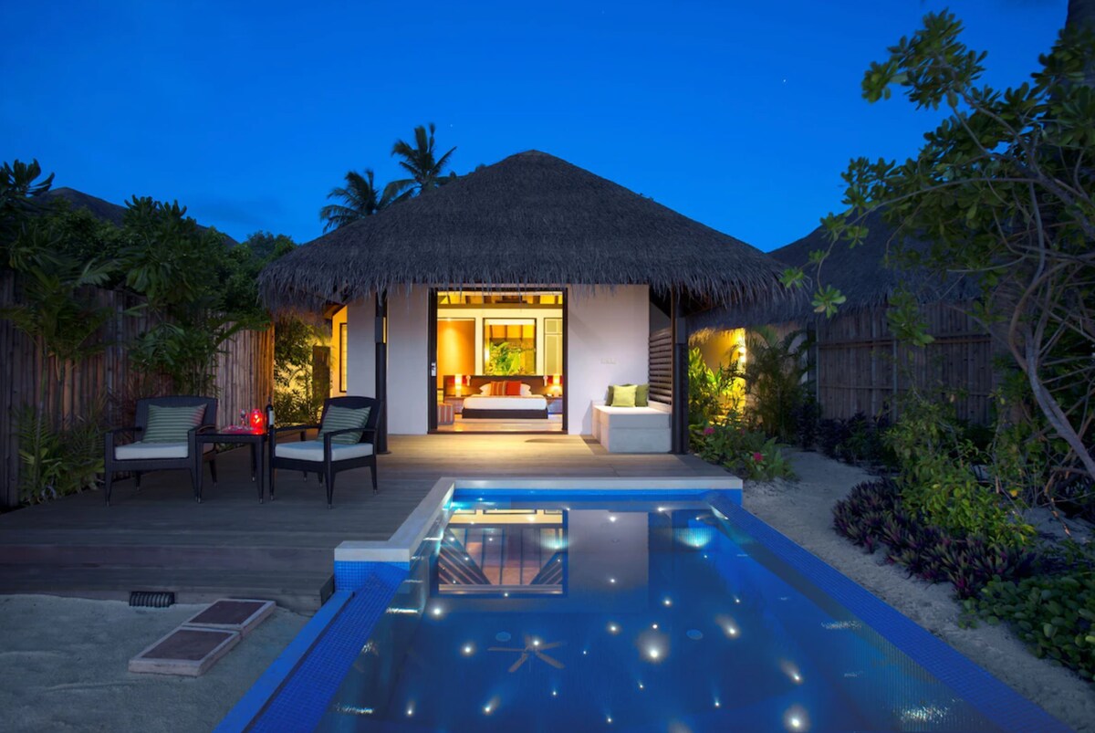 The villa features an open layout with a thatched roof, offering a view of the private pool and garden. Comfortable seating areas are arranged on a wooden deck, and ambient lighting enhances the tranquil atmosphere as evening sets in.