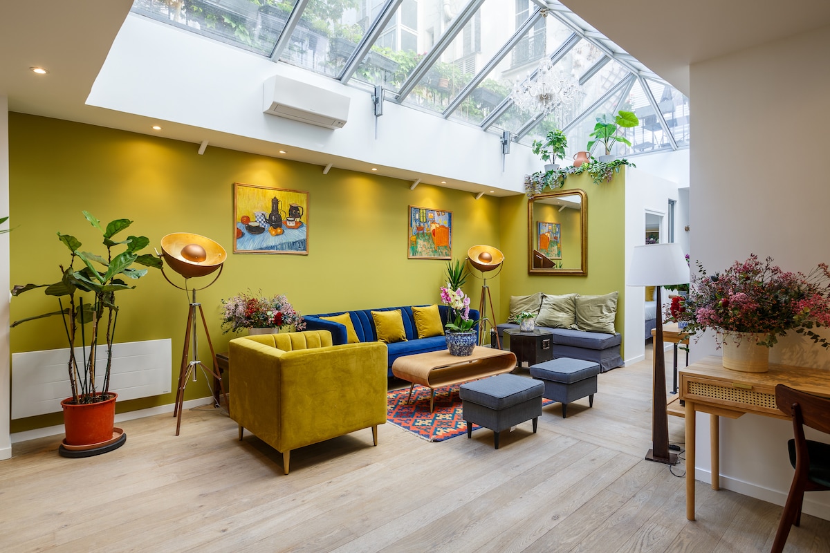 A spacious living area features a mix of seating options, including a vibrant yellow sofa and a muted gray couch. Bright plants and artwork adorn the walls, while natural light streams through a glass skylight, creating a fresh and inviting atmosphere.