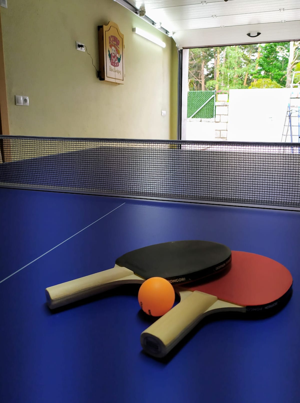 A ping pong table is set up indoors, featuring a blue playing surface with a net in place. Two paddles, one red and one black, and an orange ping pong ball are positioned on the table, ready for use.
