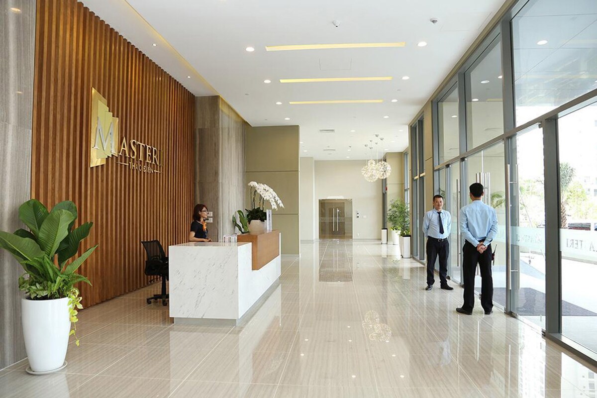 A spacious lobby is highlighted by soft natural lighting and elegant flooring. A reception desk with a welcoming staff member is positioned against a wood-paneled feature wall. Lush green plants are placed in white pots, adding a touch of tranquility to the modern space.
