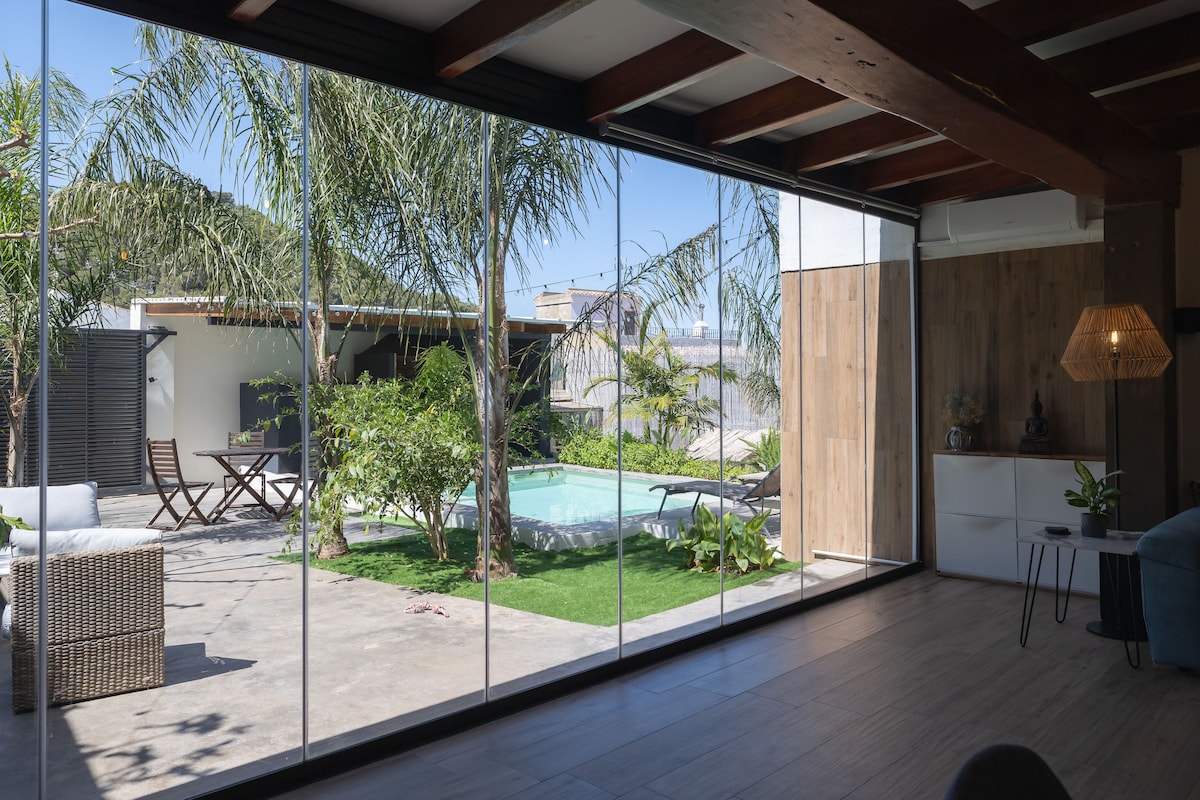 A spacious living area features expansive glass doors that open to a well-maintained garden. A turquoise pool is visible amid tropical plants, with a seating area on the patio. Natural light floods the interior, enhancing the serene connection between indoor and outdoor spaces.