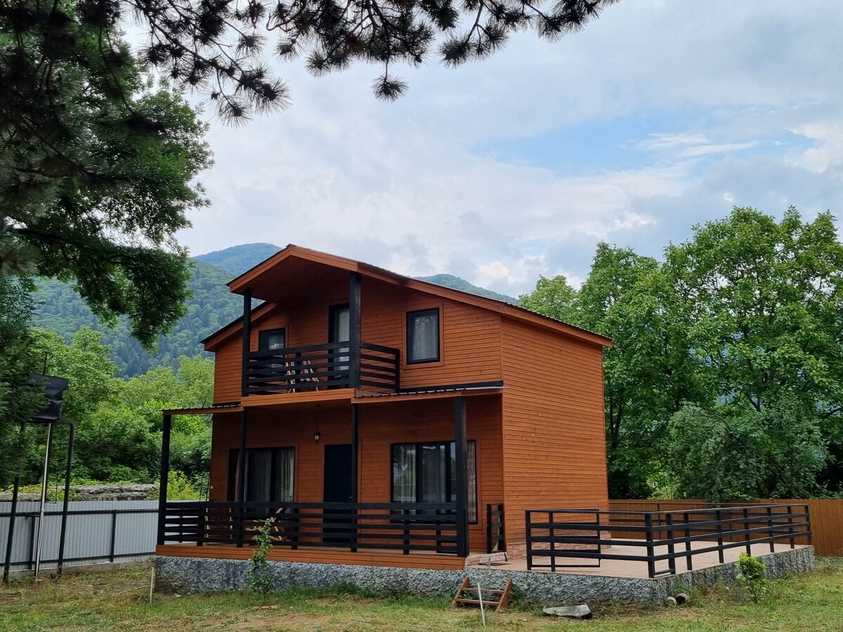 A two-story cottage made of natural wood features a spacious front patio surrounded by greenery. Dual balconies provide access to upper rooms, while large windows invite natural light into the living spaces. The backdrop includes lush trees and rolling mountains.