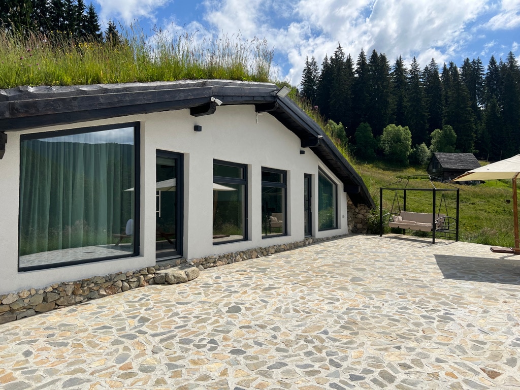 A modern bungalow features large windows that provide ample natural light and views of the surrounding greenery. The exterior showcases a stone-paved area, complemented by a grassy roof and a backdrop of trees. A swing set is visible, enhancing the outdoor space.