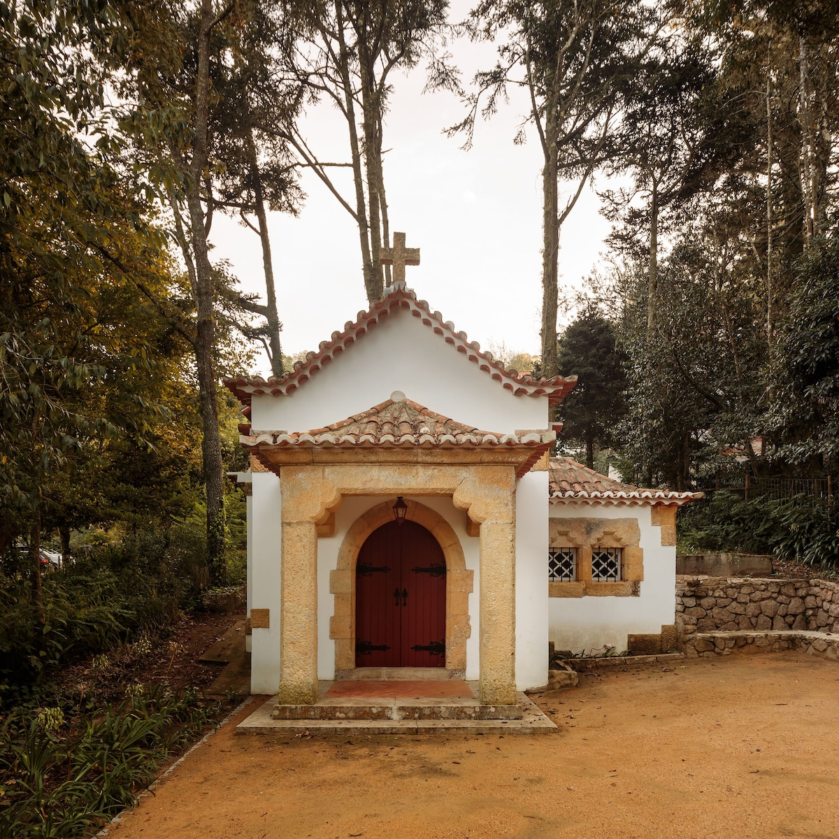 A century-old chapel is nestled among trees, showcasing traditional architecture with a red door and a distinctive cross. The white exterior features stone accents, while a tiled roof complements the natural surroundings, creating a serene and historical setting.