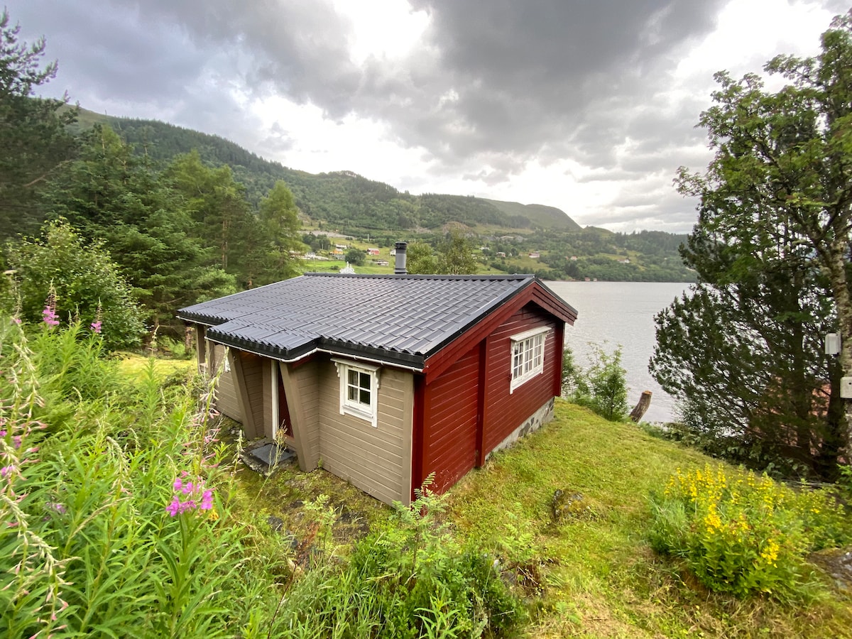 A colorful cabin is nestled amidst greenery, featuring a blend of red and gray exterior. A tiled roof provides shelter. The surrounding landscape includes wildflowers and trees, with a view of the serene body of water nearby and mountains in the background, under a cloudy sky.