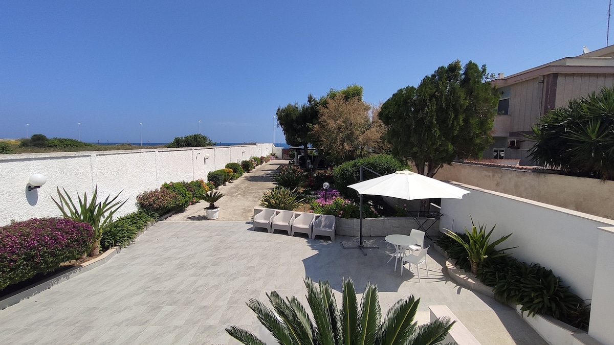 A spacious garden area is seen, featuring a variety of Mediterranean plants and flowers. Lounge chairs provide seating options under a white umbrella. A paved pathway leads towards a backdrop of trees and shrubs, enhancing the natural surroundings of the property.