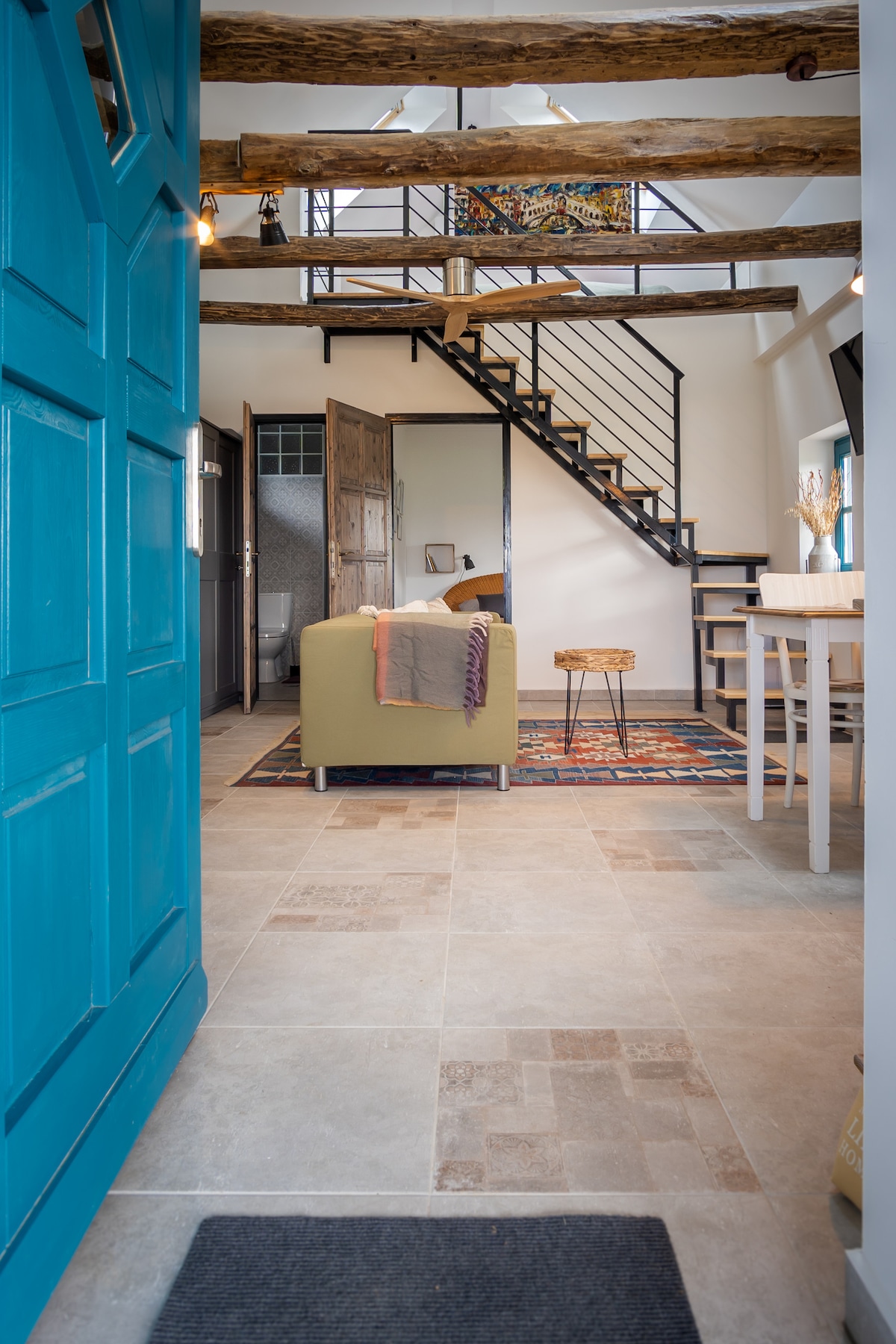 A welcoming interior is visible, featuring a bright blue door leading to a spacious living area. Natural wooden beams cross the ceiling, enhancing the rustic charm. A cozy green seating arrangement is complemented by a decorative rug and a small table, creating an inviting space.