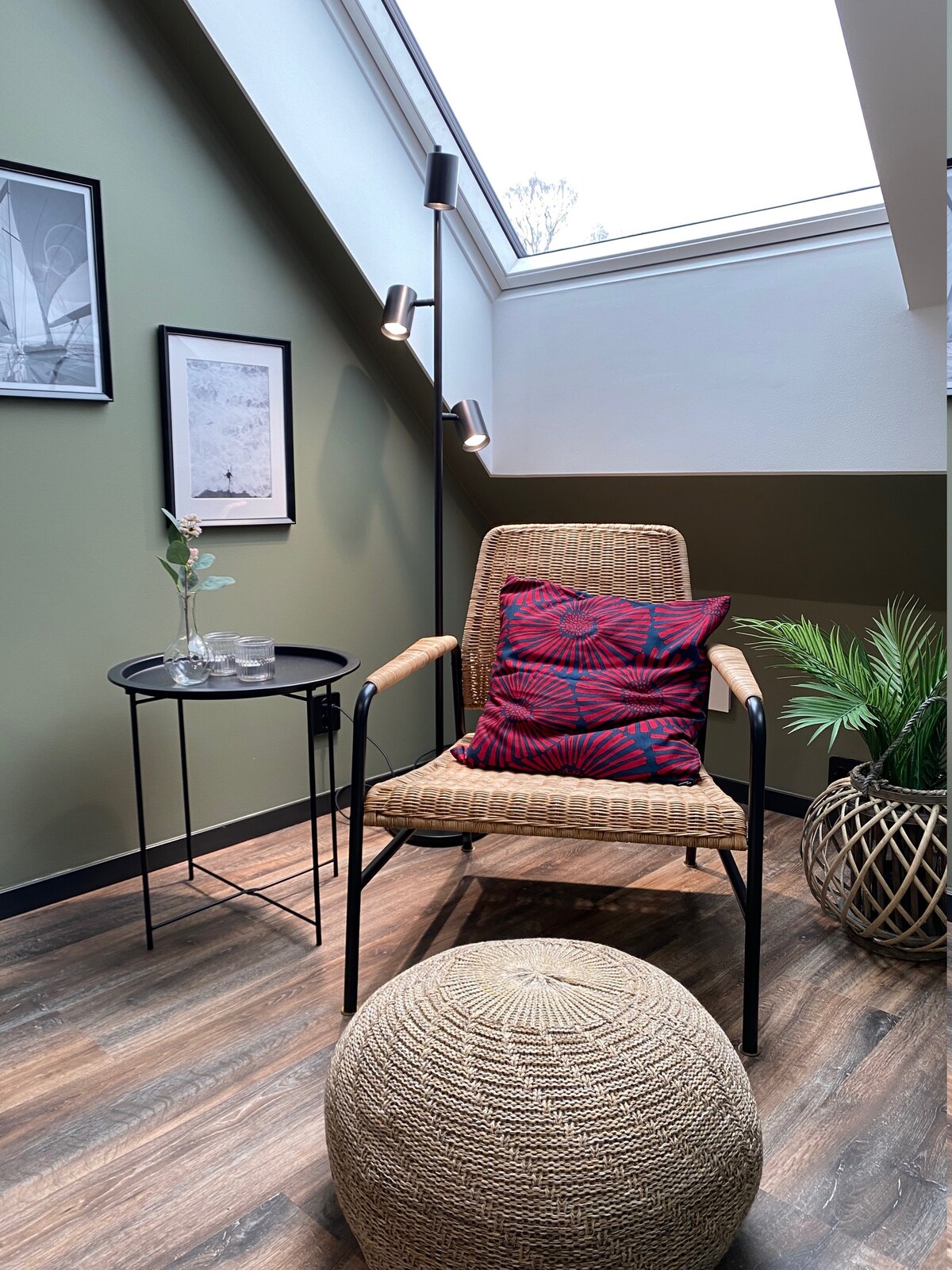 A cozy seating area is showcased with a woven chair adorned with decorative cushions. A small round table stands beside it, featuring glassware. A soft, natural fiber pouf is placed on the floor, while a potted plant adds a touch of greenery. Natural light enters through a skylight.