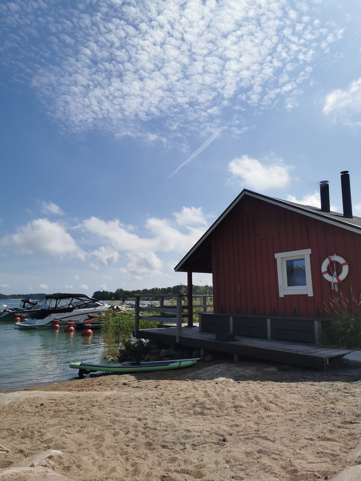 A red wooden cabin is positioned near the water's edge, featuring a small porch with steps leading down to the sandy area. A life preserver hangs on the wall, and a few boats can be seen anchored nearby. The sky is largely clear with scattered clouds.