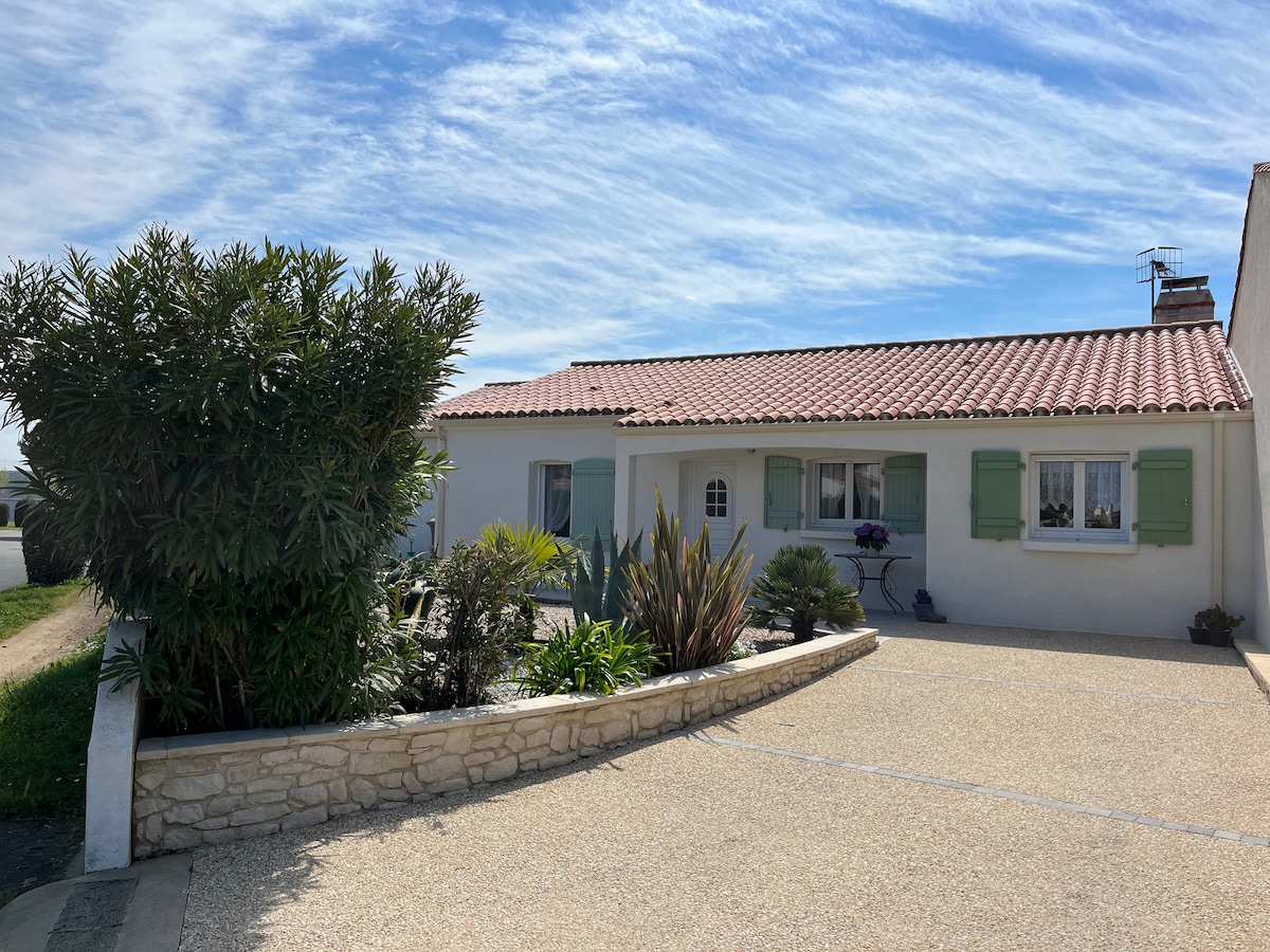 The exterior of a single-story house is shown, featuring a red-tiled roof and green shutters. A landscaped garden with various plants enhances the curb appeal. A gravel pathway leads to the entrance, bordered by a low stone wall.