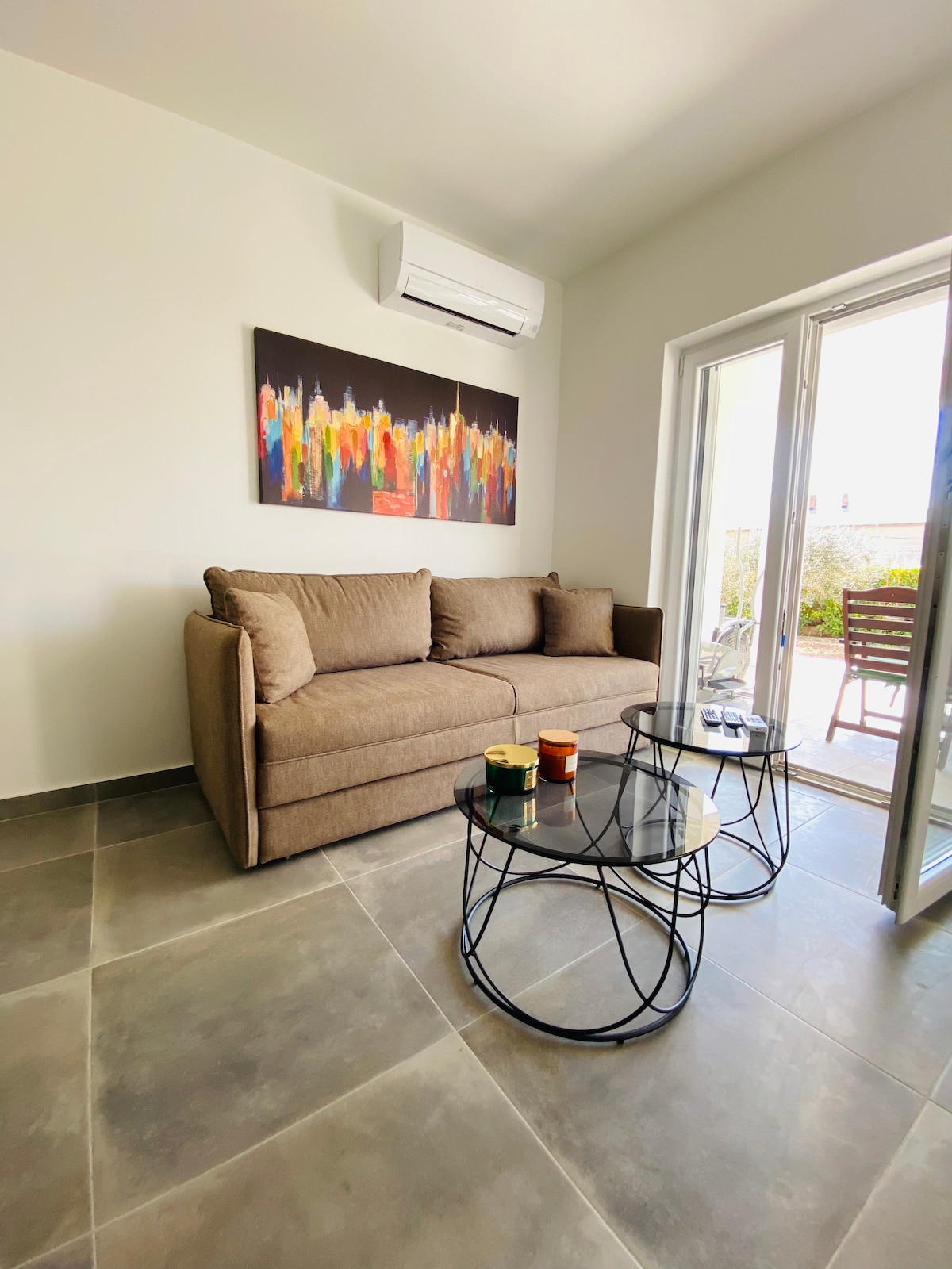 A comfortable seating area includes a light brown sofa positioned near a large window. Two circular glass-topped tables are nearby, placed on a tiled floor. A colorful wall art piece adds visual interest to the neutral wall, while natural light fills the space.