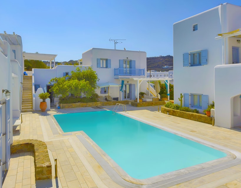 A clear blue pool is surrounded by beige stone paving, with lounge chairs placed around its perimeter. White buildings with light blue shutters are visible in the background, and a few potted plants add greenery to the scene. A staircase leads to an upper level in the structure.