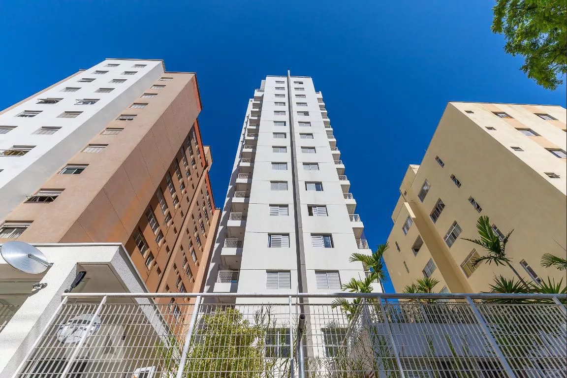 A modern multi-story building is surrounded by neighboring structures. The exterior features a blend of light and darker hues, and the upper floors appear to offer ample natural light. Lush greenery can be seen at the base, contributing to the urban atmosphere.