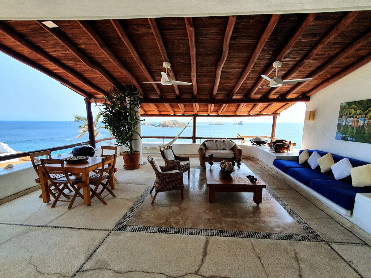 An open-air terrace features a spacious seating area with a blue sofa and wicker chairs. A wooden table is surrounded by four chairs. The stunning view of the ocean is framed by wooden beams and ceiling fans, enhancing the relaxed atmosphere.