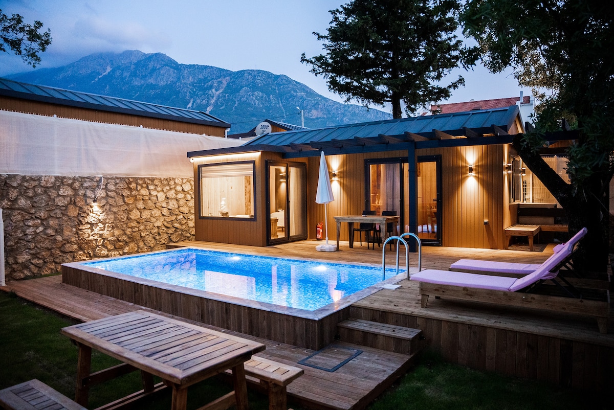 A modern bungalow is depicted with a private swimming pool surrounded by wooden decking. Comfortable lounge chairs provide space for relaxation. Soft lighting casts a warm glow over the stone walls and patio area, while distant mountains are visible, enhancing the serene setting.
